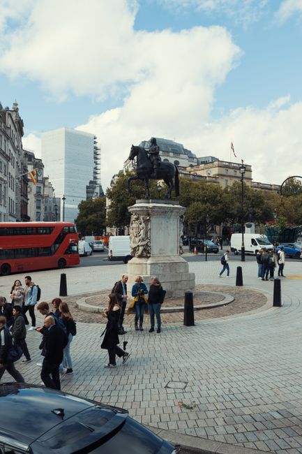 A bustling city square in Trafalgar Square, featuring a large stone fountain with water cascading from its multiple tiers in the foreground, surrounded by historic architecture including a white class
