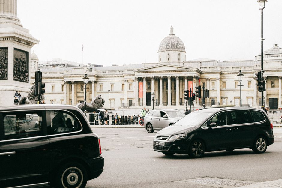 A busy urban square in London featuring the bronze statue of a mounted figure on a stone pedestal at the center. Surrounding the monument is a paved circular area with black bollards marking its perim